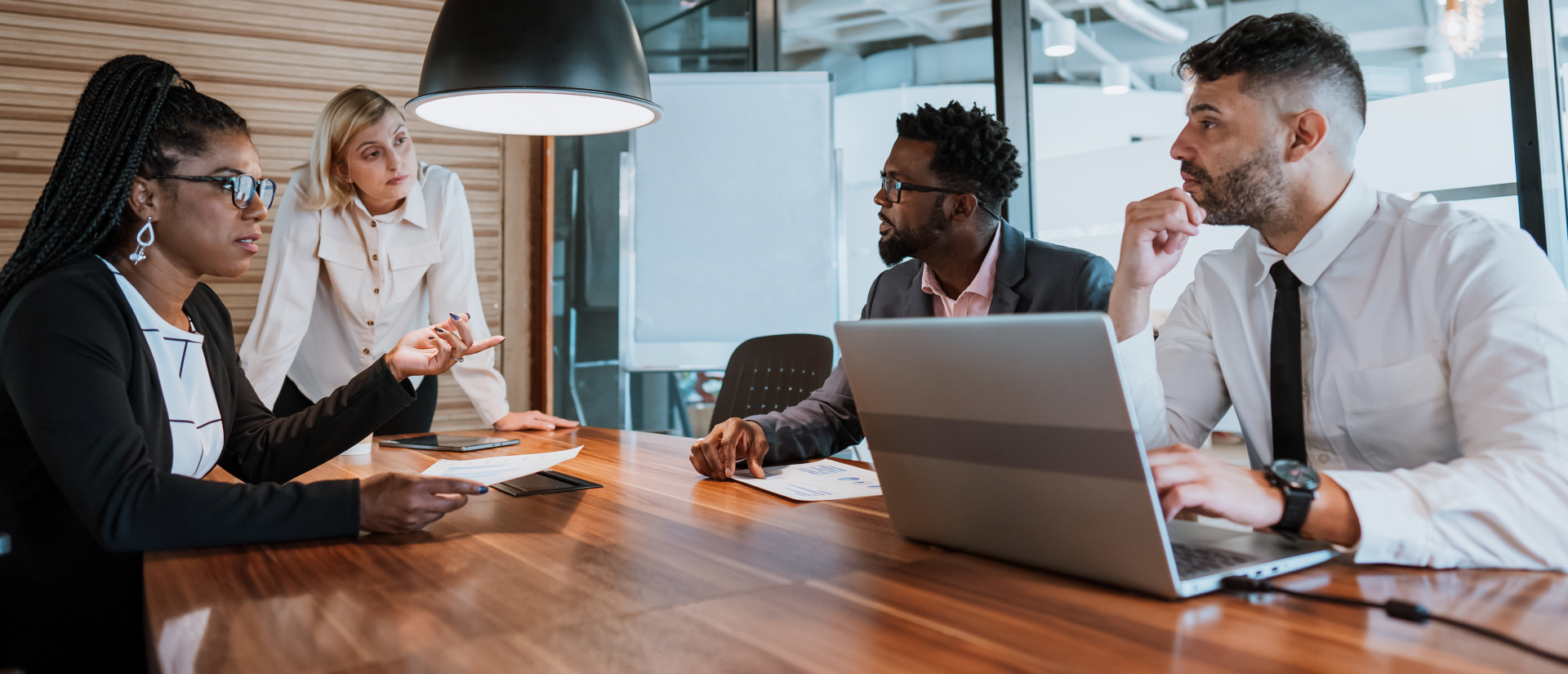 Four professionals engaged in a team meeting around a wooden conference table with laptops and documents.