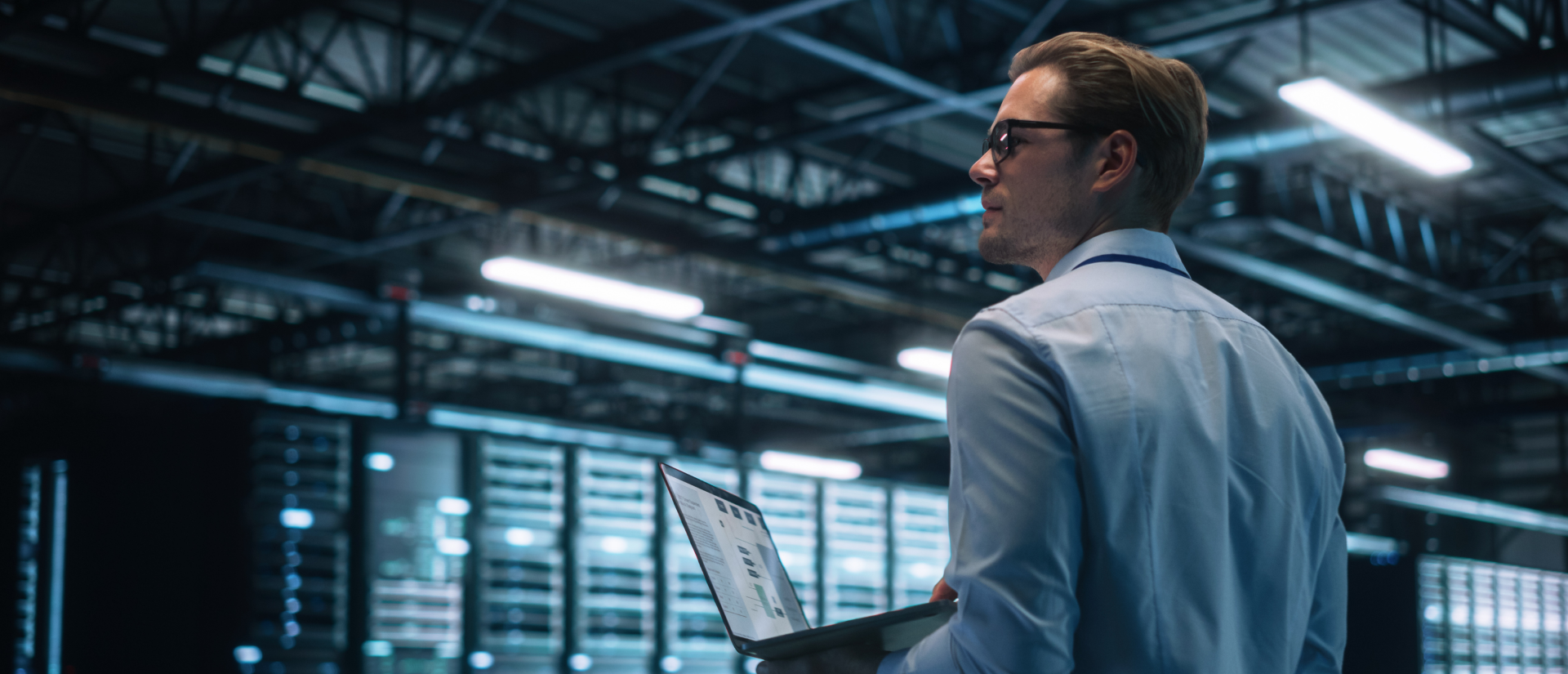 IT professional in a server facility holding a laptop and inspecting network racks and infrastructure.